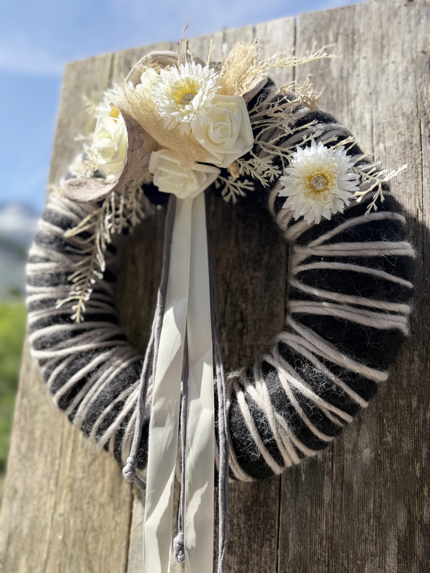 Schwarz-weißer Wollkranz mit weißen Rosen, Gerbera, Trockenblumen und weißem Satinband vor Lechtaler Bergpanorama – Naturdekoration NaturTraum Lechtal