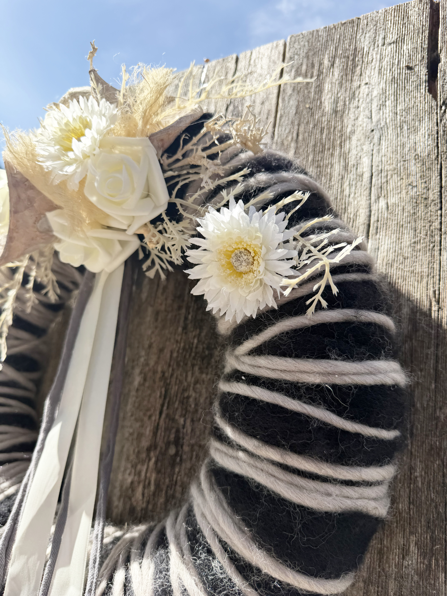 Seitenansicht schwarz-weißer Wollkranz mit weißer Gerbera, Rose und Trockenblumen vor blauem Himmel – Naturdekoration NaturTraum Lechtal