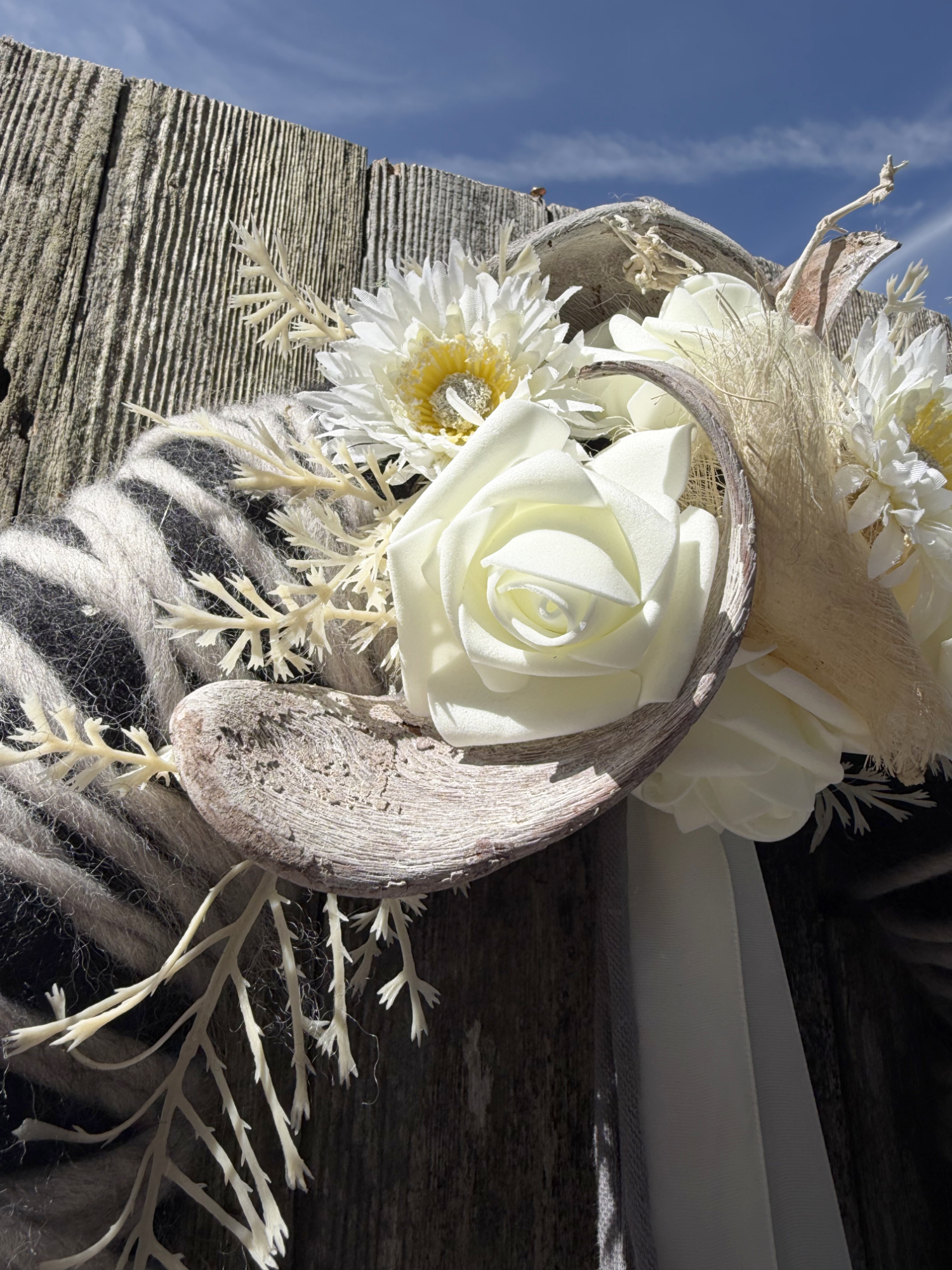Alternativtext: Nahaufnahme schwarz-weißer Wollkranz mit weißen Rosen, Gerbera und getrockneten Naturmaterialien vor blauem Himmel – Naturdekoration NaturTraum Lechtal