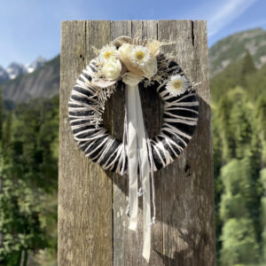 Schwarz-weißer Wollkranz mit weißen Rosen, Gerbera, Trockenblumen und weißem Satinband vor Lechtaler Bergpanorama – Naturdekoration NaturTraum Lechtal