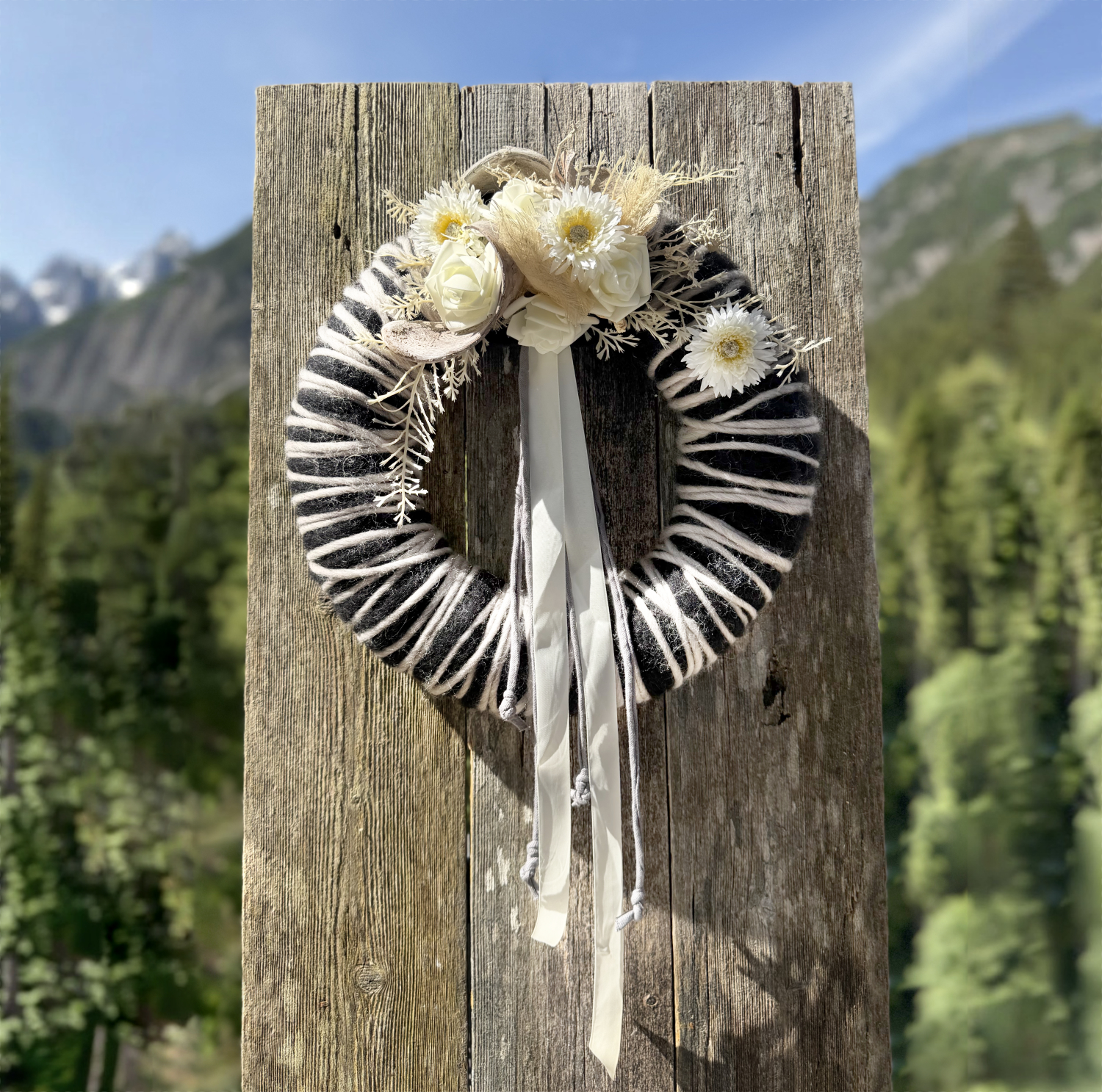 Schwarz-weißer Wollkranz mit weißen Rosen, Gerbera, Trockenblumen und weißem Satinband vor Lechtaler Bergpanorama – Naturdekoration NaturTraum Lechtal
