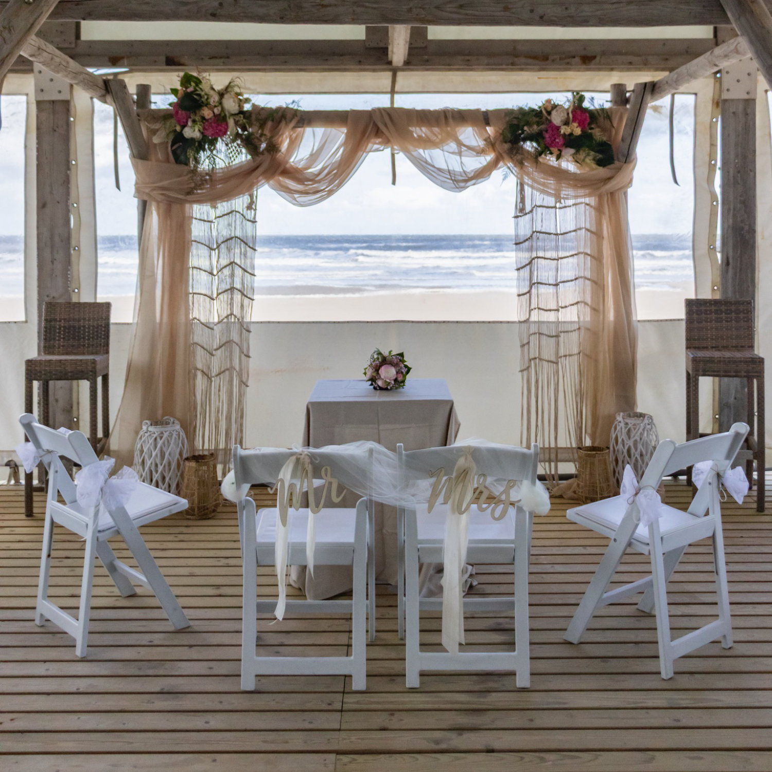 Festlich dekorierter Trauungsbereich am Meer mit Blumengestecken, goldenen Stoffbahnen, Mr & Mrs Schildern und weißen Stühlen – Hochzeitsfloristik von NaturTraum
