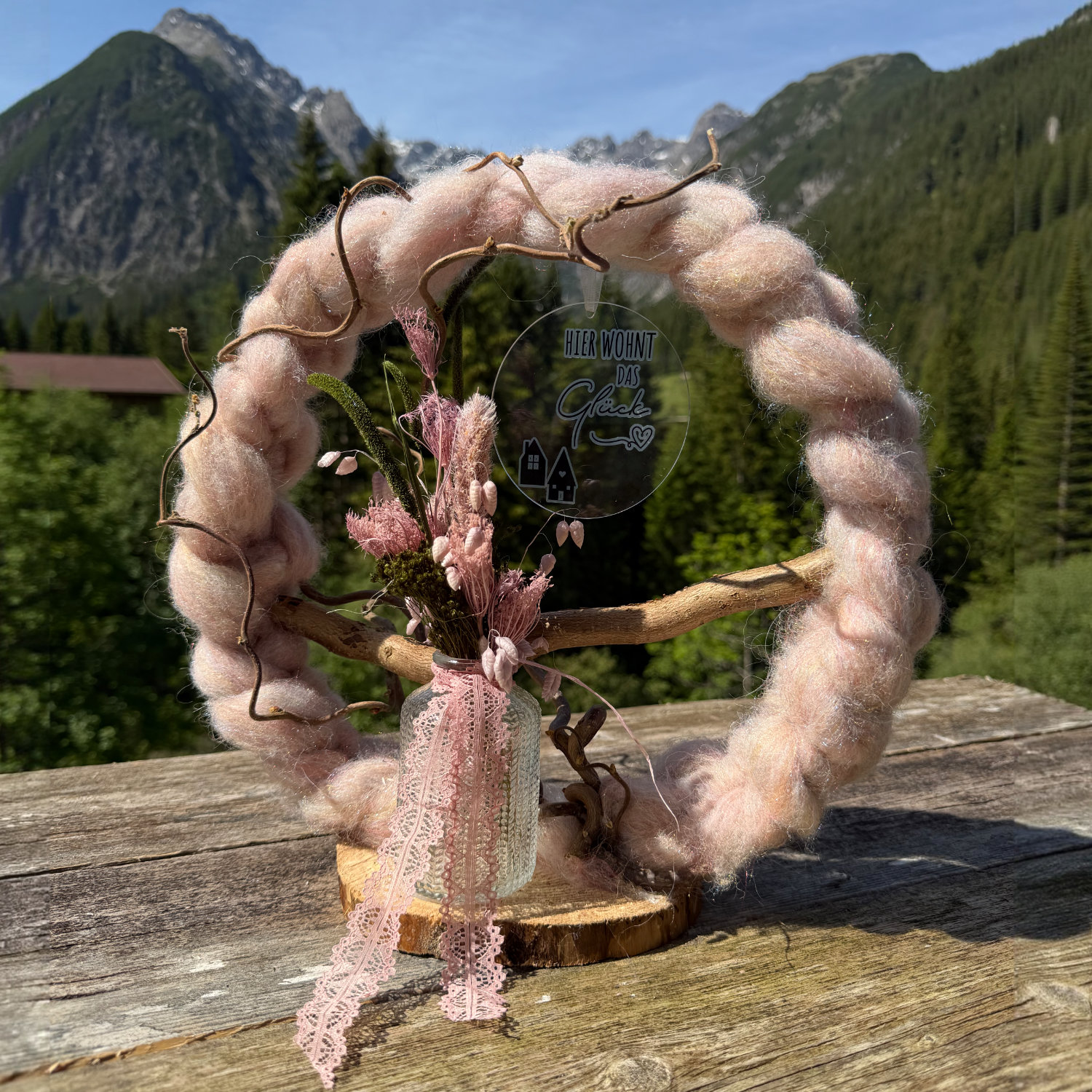 Rosafarbener Wollkranz auf Holzscheibe mit kleinem Trockenblumenstrauß, Spitzenband und Anhänger "Hier wohnt das Glück" vor Lechtaler Bergpanorama – Naturdekoration NaturTraum