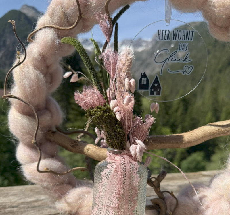 Nahaufnahme rosa Trockenblumenstrauß in Glasvase mit Spitzenband und Anhänger "Hier wohnt das Glück" an rosafarbenem Wollkranz vor Lechtaler Bergpanorama – NaturTraum