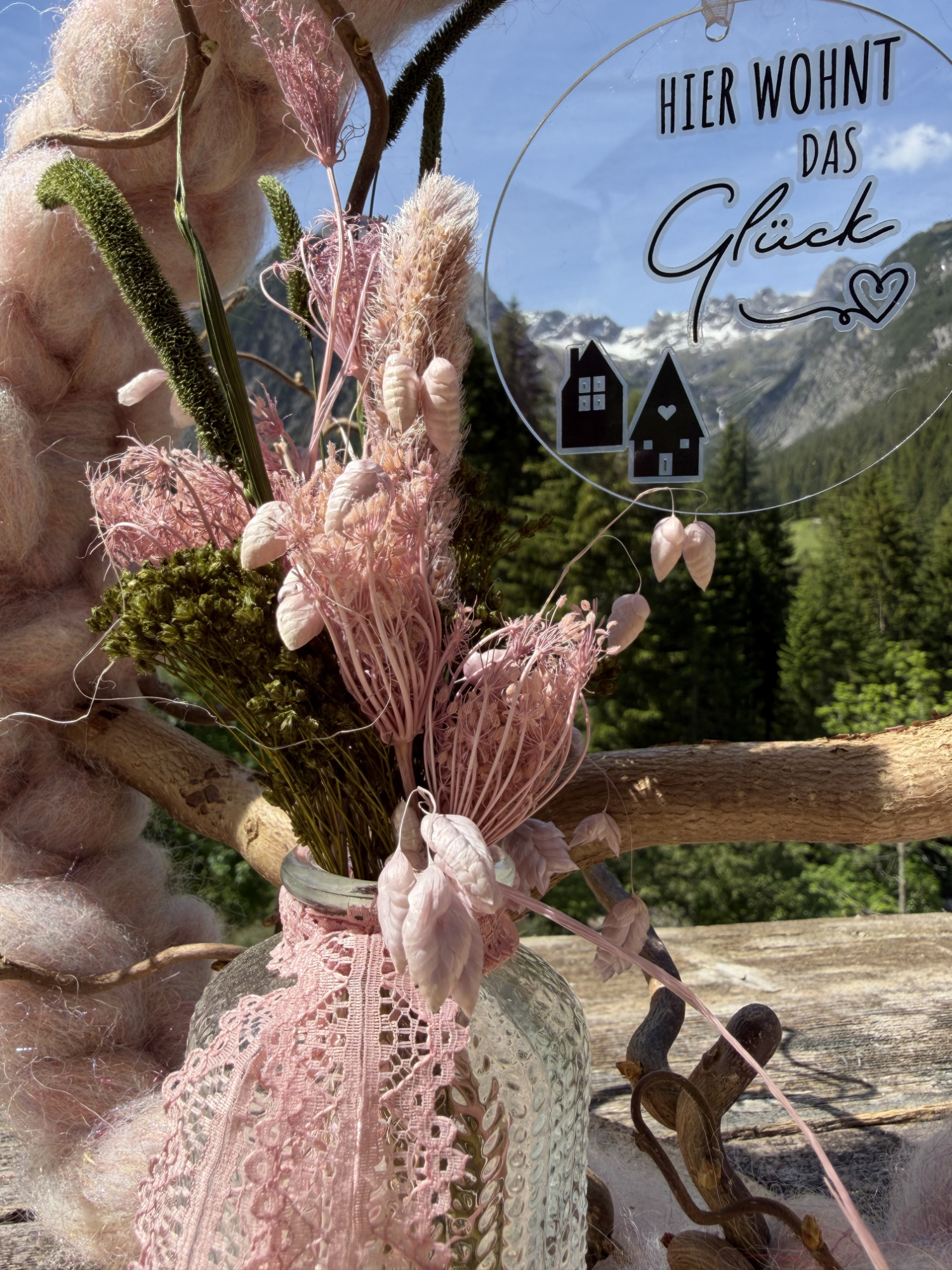 Nahaufnahme rosa Trockenblumenstrauß mit Spitzenband in Glasvase und Anhänger "Hier wohnt das Glück" vor schneebedecktem Lechtaler Bergpanorama – NaturTraum