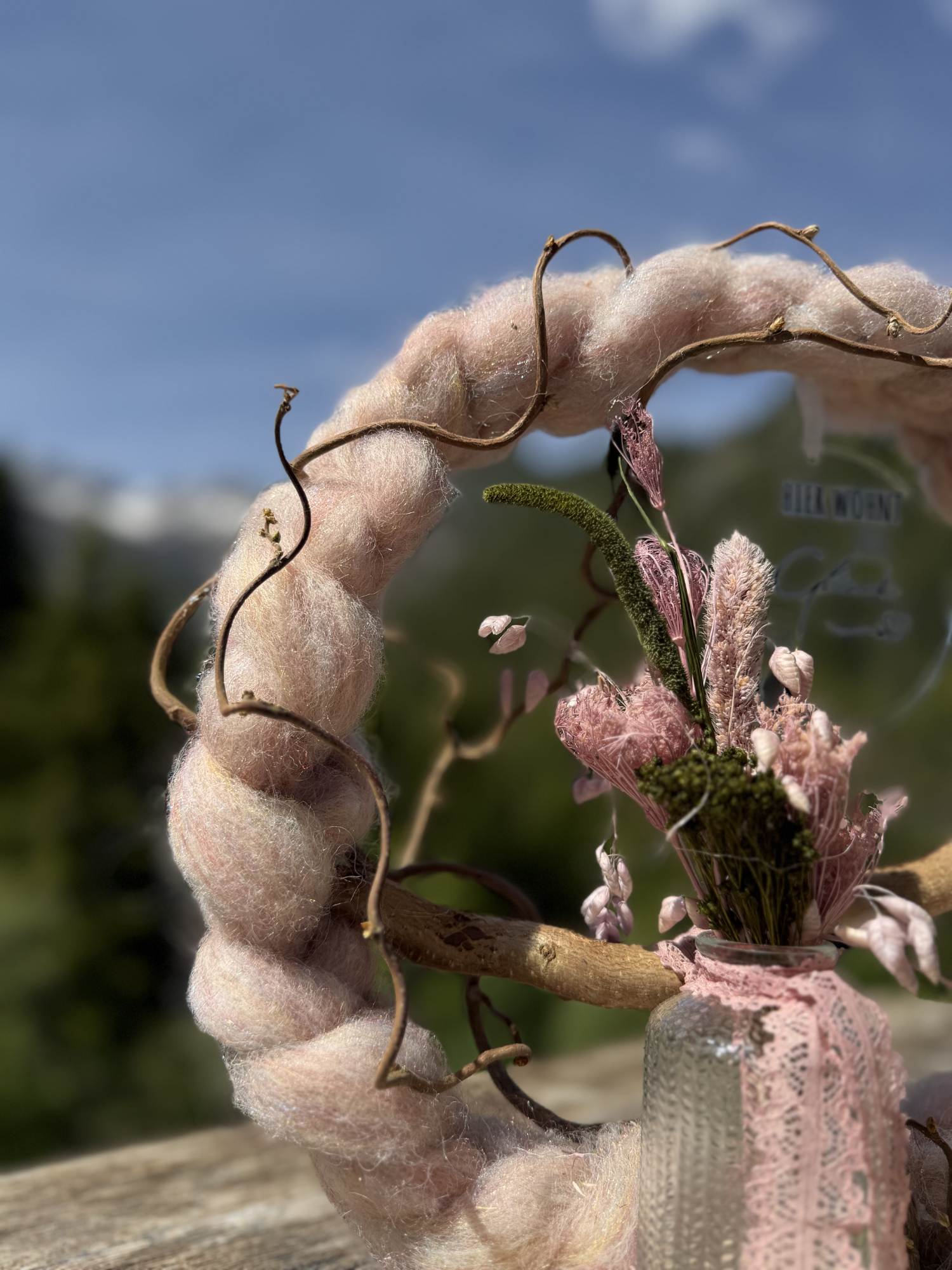 Seitenansicht rosafarbener Wollkranz mit gedrehten Ästen und Trockenblumenstrauß in Spitzenvase vor Lechtaler Bergpanorama – Naturdekoration NaturTraum