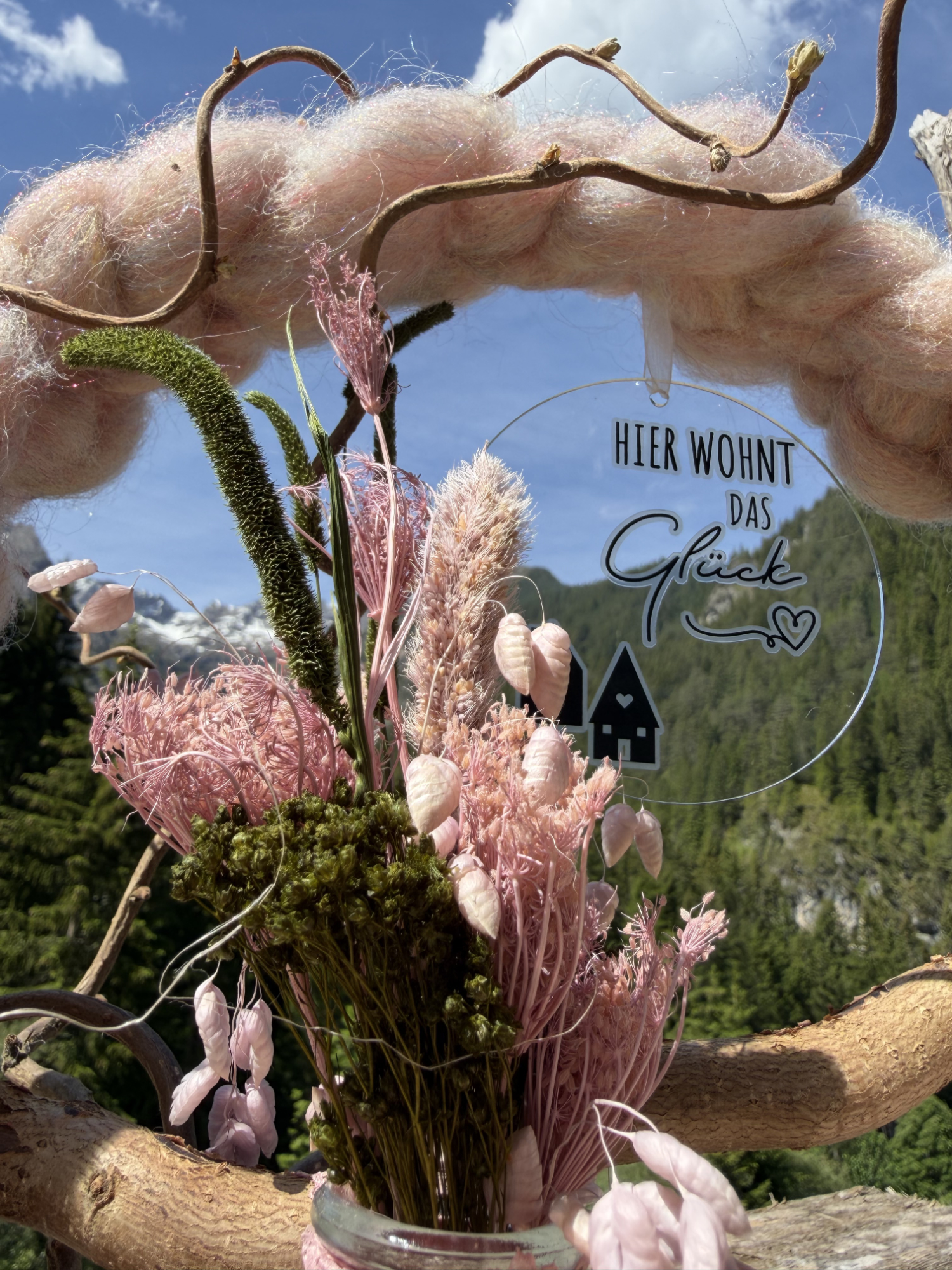 Nahaufnahme rosa Trockenblumenstrauß mit Schleierkraut, Federgras und Anhänger "Hier wohnt das Glück" an rosafarbenem Wollkranz vor Lechtaler Bergpanorama – NaturTraum