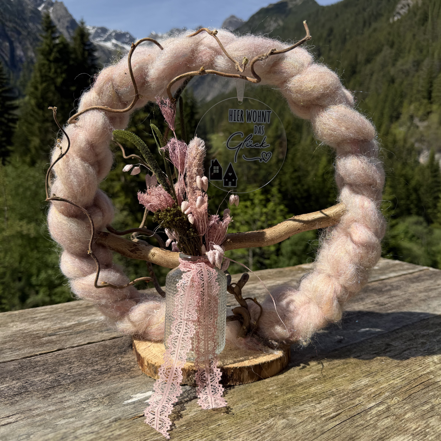 Rosafarbener Wollkranz auf Holzscheibe mit rosa Trockenblumenstrauß, Spitzenband und Anhänger "Hier wohnt das Glück" vor Lechtaler Bergpanorama – Naturdekoration NaturTraum