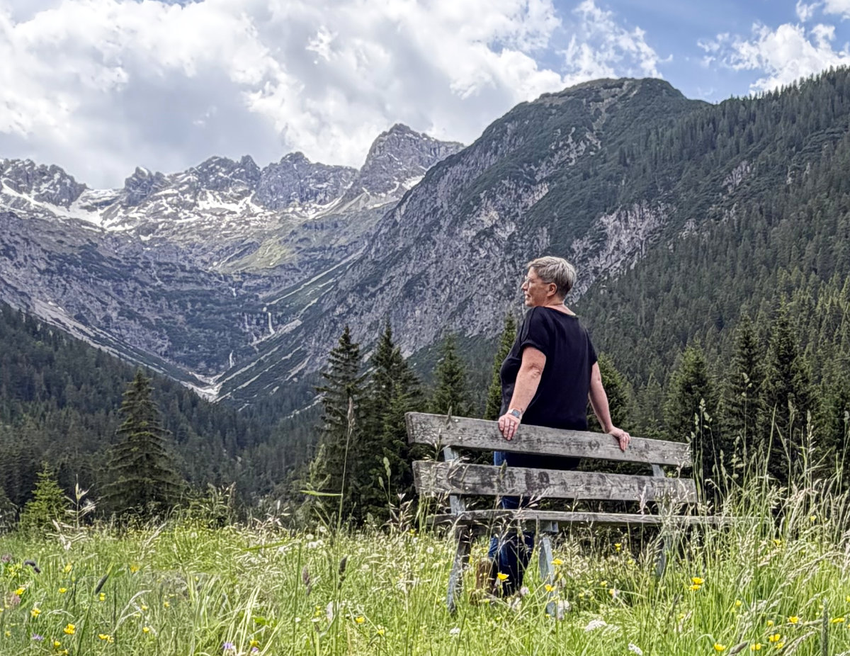 Constanze Wolf, Floristin und Inhaberin von NaturTraum, sitzt auf einer Holzbank in einer Bergwiese und blickt ins Lechtaler Bergpanorama – Gramais, Tirol