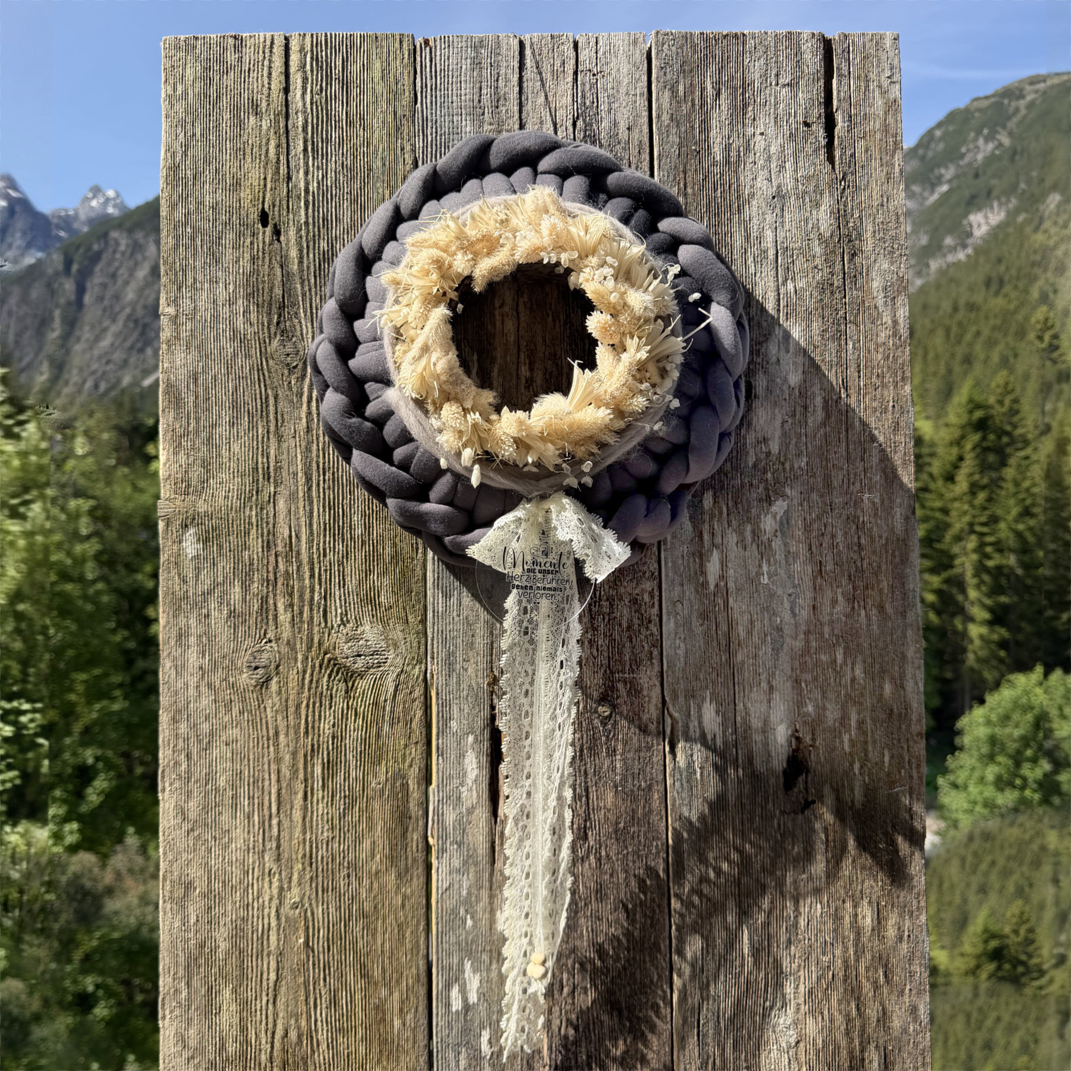 Dunkelgrauer Stoffkranz mit cremefarbenen Trockenblumen, Spitzenband und Anhänger "Momente die berühren gehen niemals verloren" vor Lechtaler Bergpanorama – Naturdekoration NaturTraum Lechtal